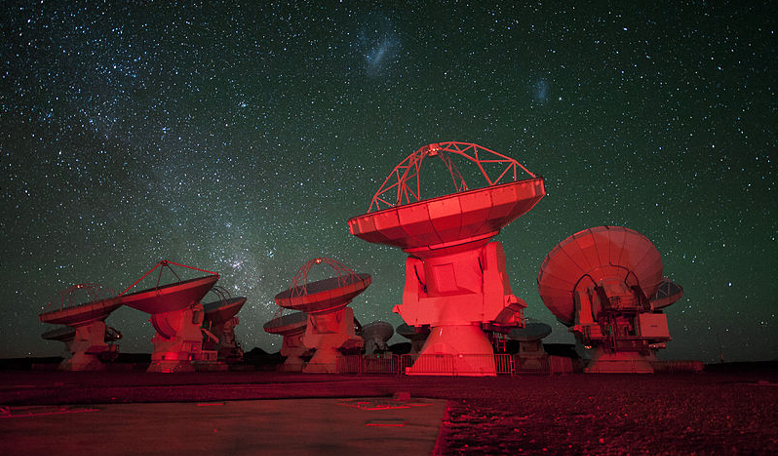 ALMA antennae bathed in red light with the southern Milky Way on the left in the background and the Magellanic Clouds at the top above the main antennae. Image: ESO/C. Malin