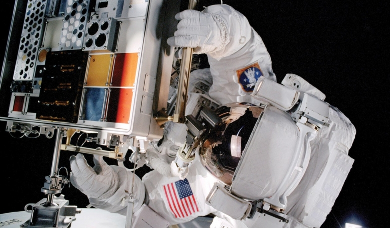 Spacewalking astronaut installing a tray supporting numerous material samples at the International Space Station.
